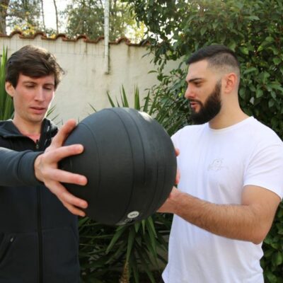 Personal fitness trainer coaching a client during a standing medicine ball exercise outdoors.