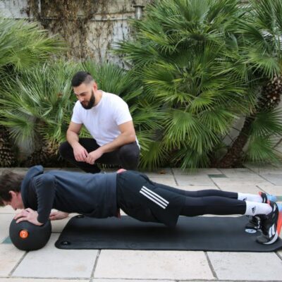 Client performing a push-up with hands on a medicine ball while a personal trainer supervises outdoors.